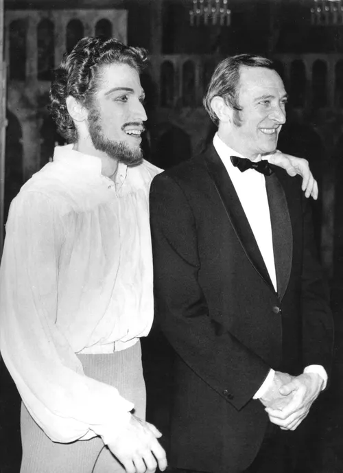 Schwarzweißfoto: Choreograf John Cranko im Smoking steht lächelnd neben einem Tänzer in historischer Bühnenkleidung mit Hemd und Bart. Der Tänzer hat den Arm freundschaftlich um Crankos Schulter gelegt. Beide wirken entspannt und fröhlich.