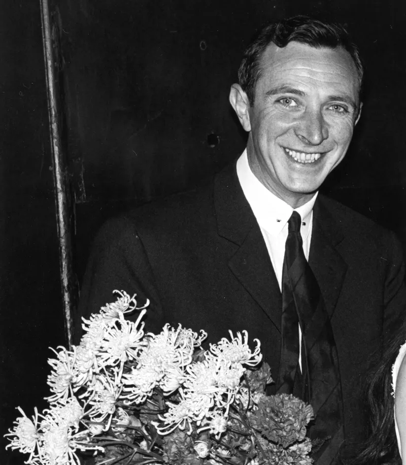 Schwarz-Weiß-Porträt des lächelnden Choreografen John Cranko. Er trägt einen Anzug und hält einen großen Strauß weißer Chrysanthemen, vermutlich nach einer erfolgreichen Ballettaufführung.